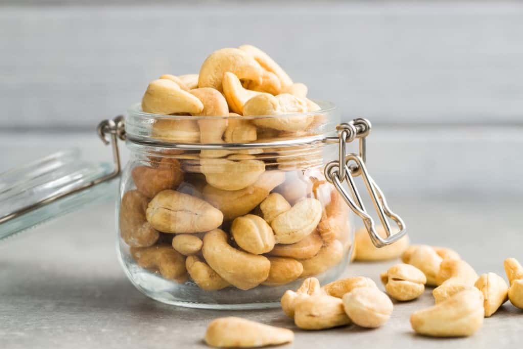Front view of small lidded clear glass jar of roasted cashew nuts on a light grey stone surface, with cashew nuts overflowing from the jar onto the surface.