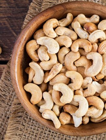 Aerial view of a wooden bowl of roasted cashew nuts placed on a burlap mat and resting on a rustic wood surface.