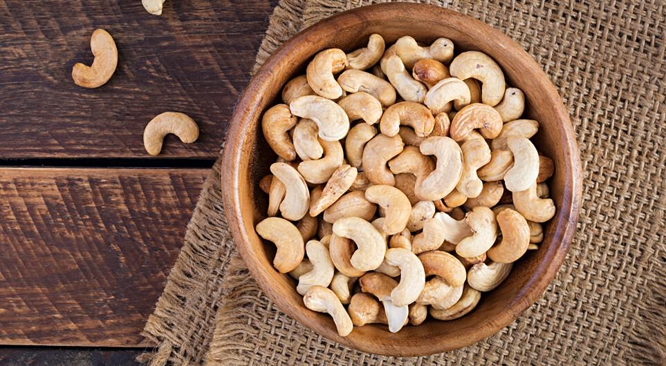 Aerial view of a wooden bowl of roasted cashew nuts placed on a burlap mat and resting on a rustic wood surface.
