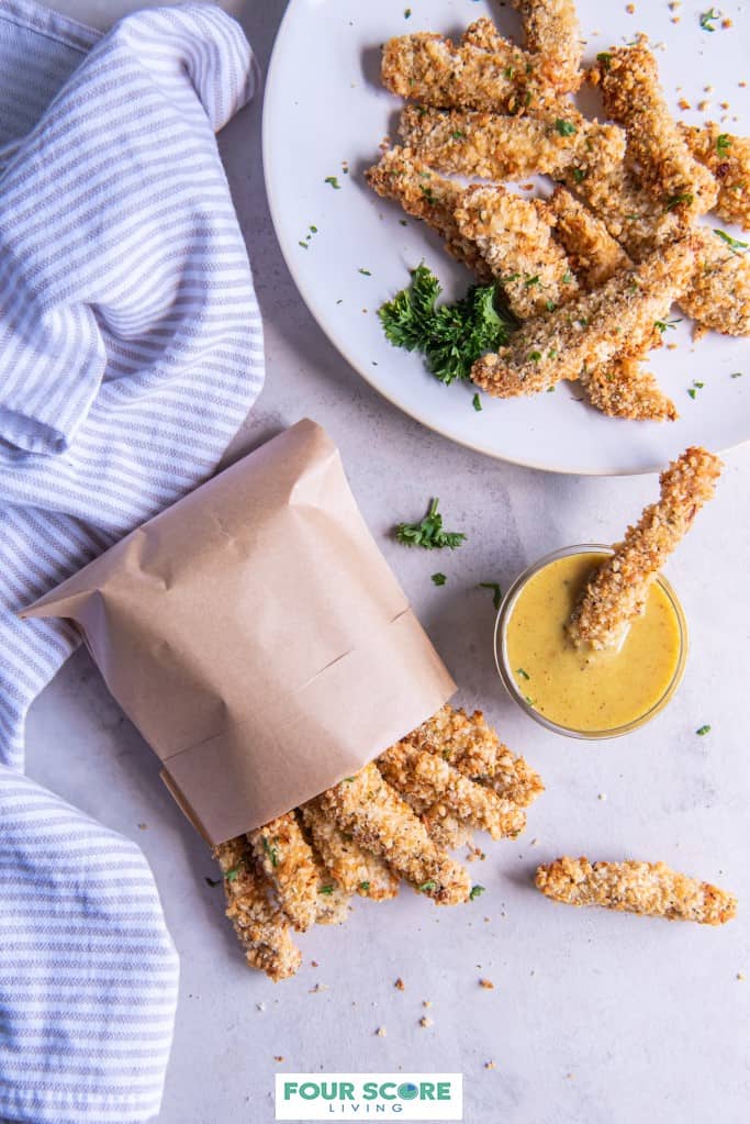 Aerial view of air fryer chicken fries on a white plate with green garnish and chicken fries in a brown paper bag and one dipping into a small dish of honey mustard sauce with a finely striped kitchen towel all on a light stone surface. 