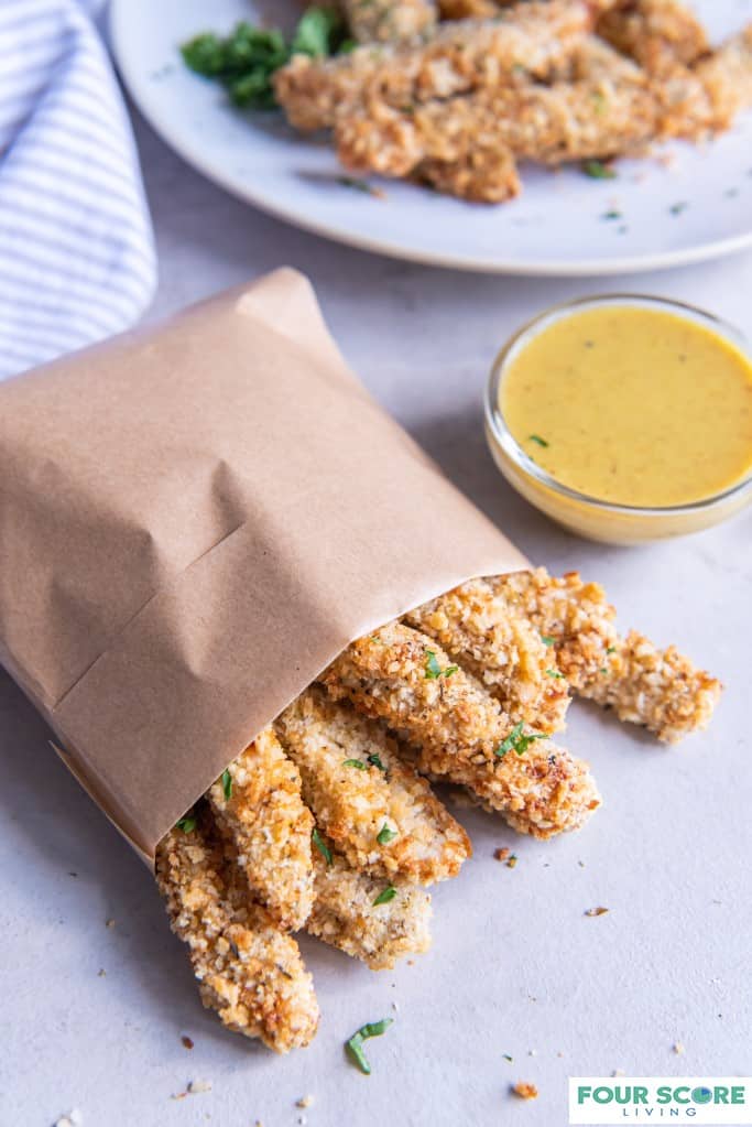 Diagonal aerial view of cooked air fryer chicken fries in a small natural brown bag with a small dish of honey mustard dipping sauce and a plate of air fryer chicken fries and a striped kitchen towel in soft focus in the background. 