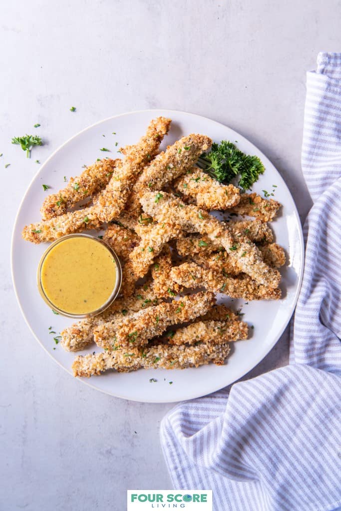 Aerial view of breaded and cooked air fryer chicken fries on a white plate with green herb garnish and a small dish of honey mustard dipping sauce with a light grey and white striped kitchen towel on a white stone surface. 