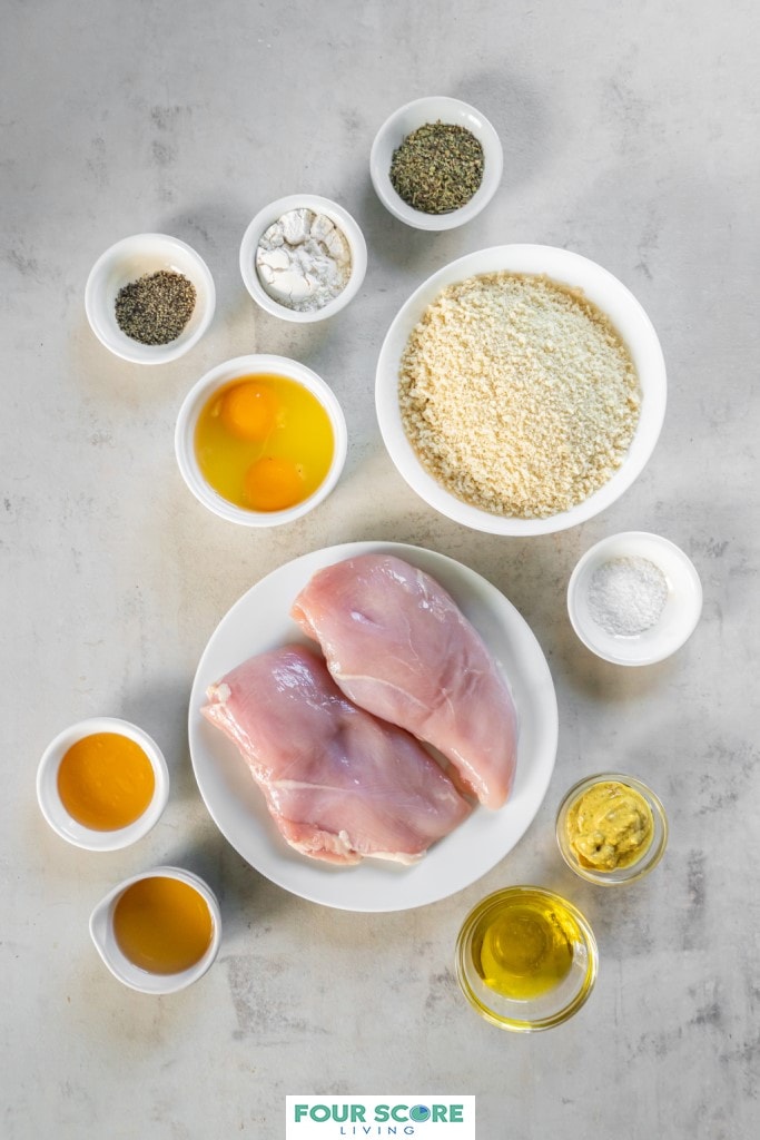 Aerial view of the ingredients in small and medium white dishes, to make air fryer chicken fries including bread crumbs, cracked raw eggs, two chicken breasts, seasonings, and flour and ingredients for honey mustard dipping sauce, honey, mustard, olive oil and apple cider vinegar. 