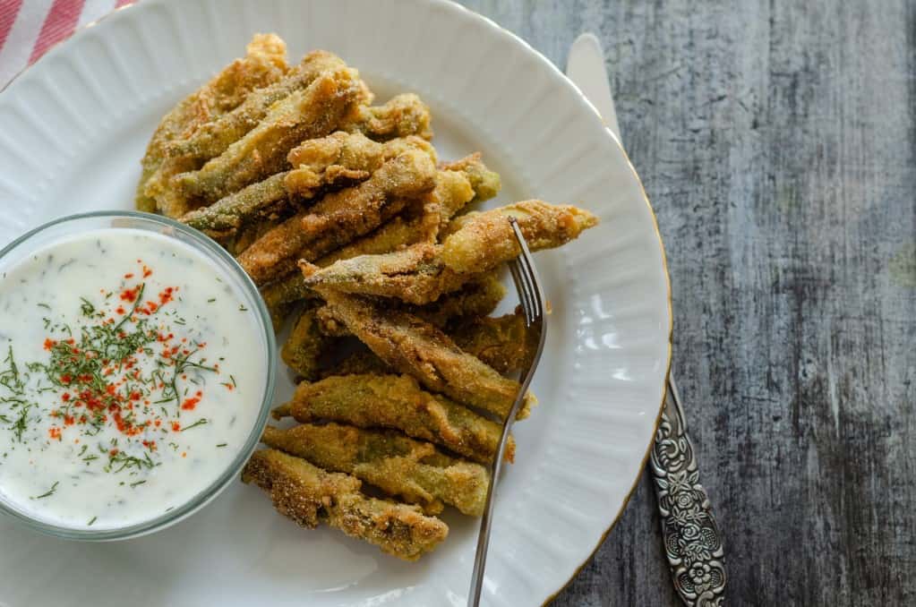 Aerial view of fried okra on a white plate with a dish of herbed cream sauce and a fork spearing an okra, resting on a rustic wood surface with a butter knife and a red and white kitchen towel in the background.
