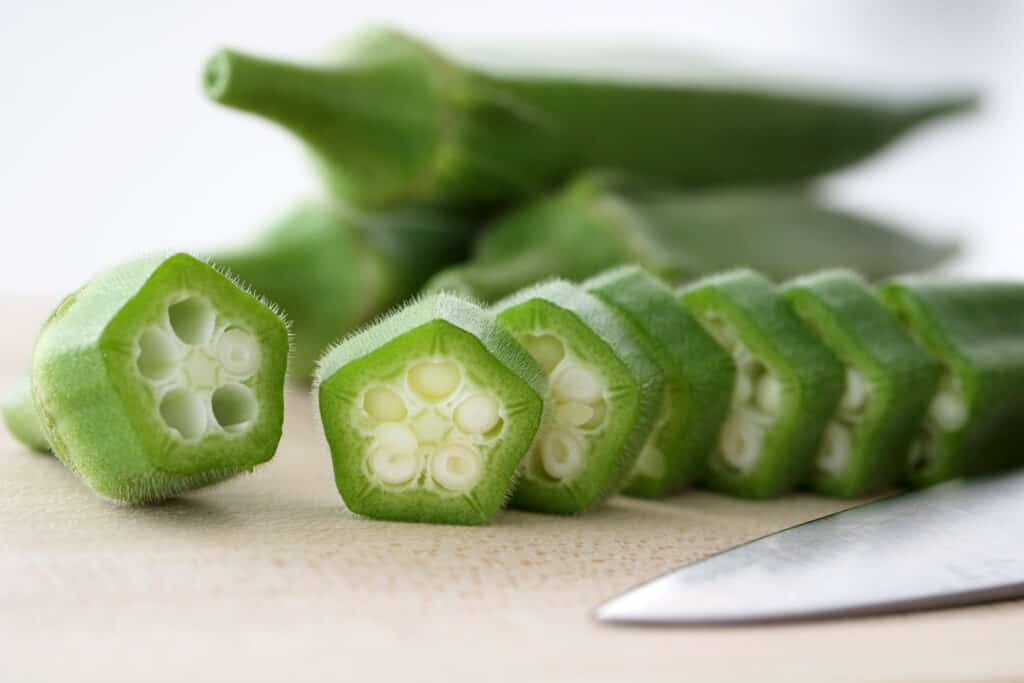 Close up frontal view of raw okra sliced into discs with a sharp knife and three whole okra in soft focus in the background all resting on a light colored wooden surface. 