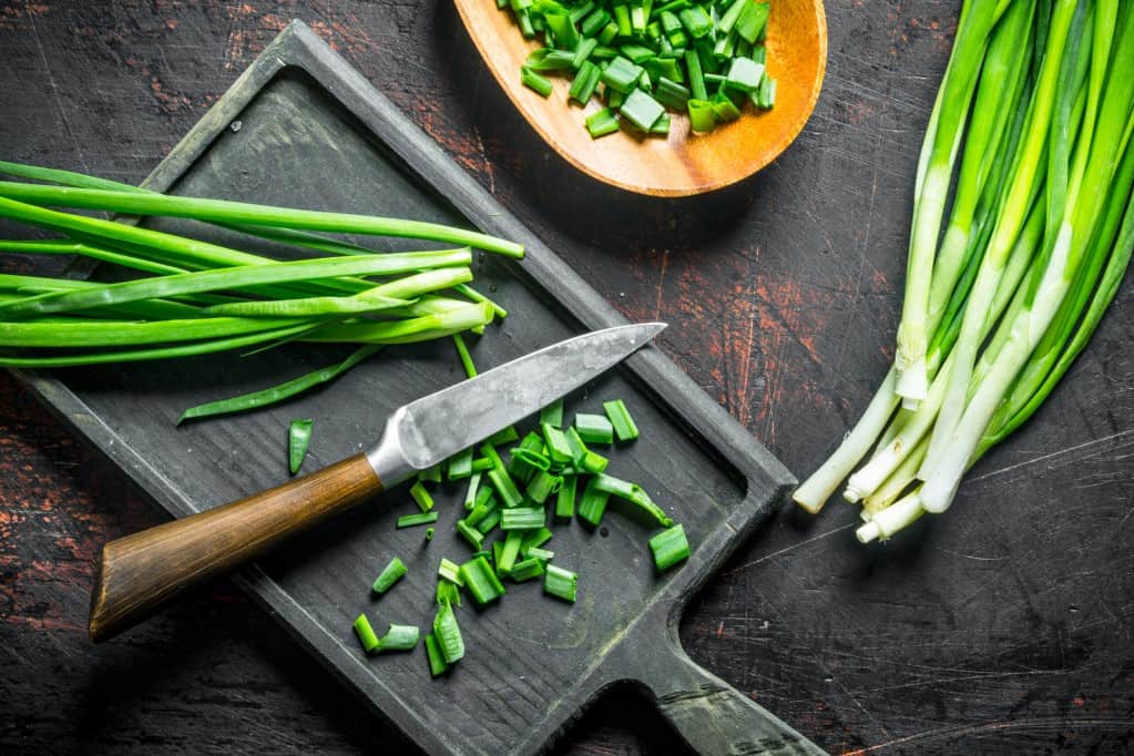 Aerial view of whole and chopped green onion on a cutting board with a shape knife and a wooden dish of chopped green onions all resting on dark rustic wood surface.
