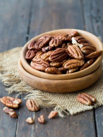 Diagonal view of a wooden bowl of raw pecans resting on a square of burlap, placed on a dark wooden, rustic surface.