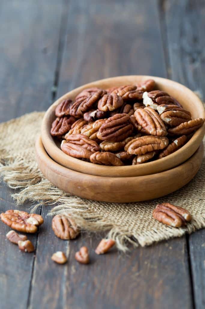 Diagonal view of a wooden bowl of raw pecans resting on a square of burlap, placed on a dark wooden, rustic surface.
