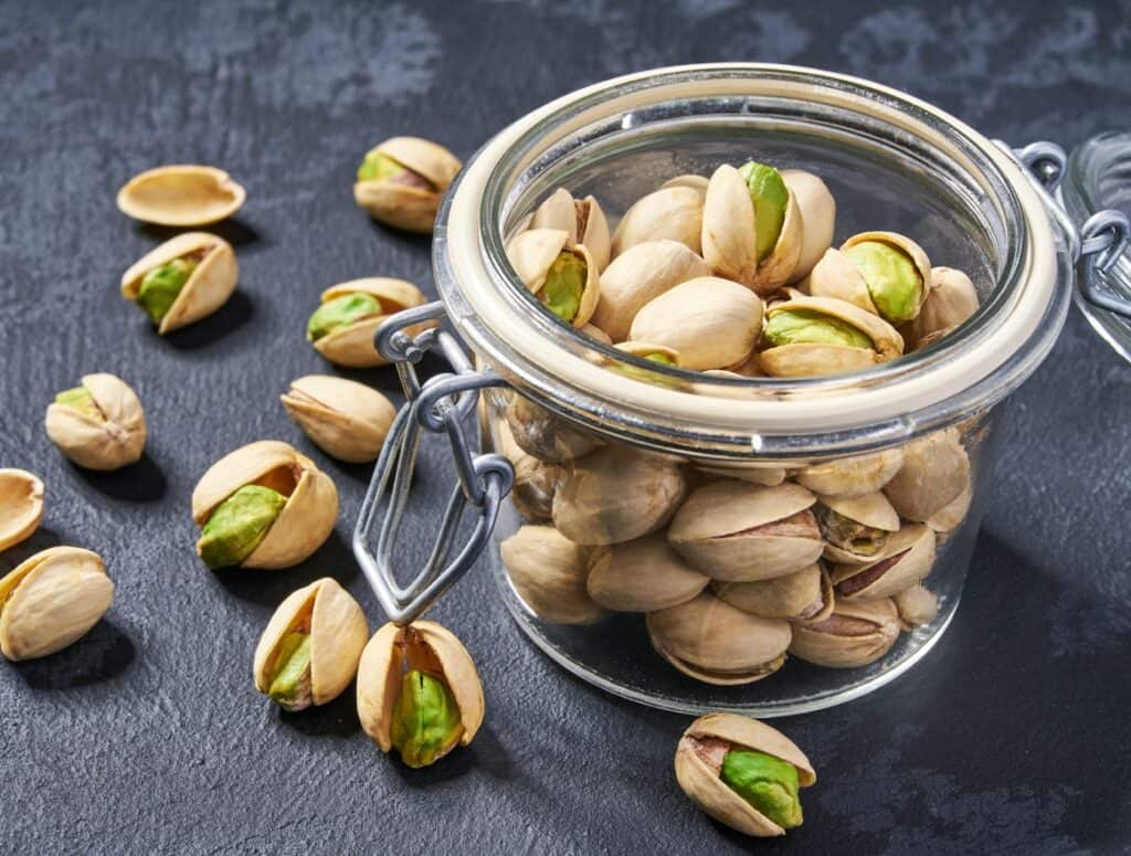 Diagonal closeup view of a hinge lidded small glass jar filled with pistachio nuts in the shell with pistachio nuts in the shell scattered on the dark slate surface.