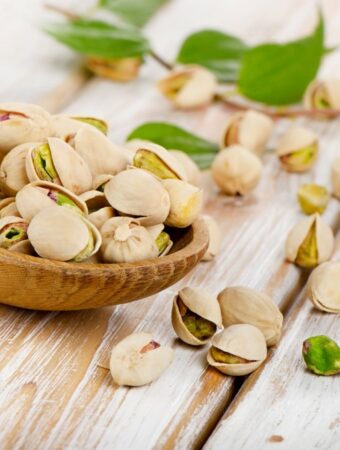 Front diagonal view of medium wooden spoon filled with pistachio nuts in the shell, with pistachios scattered with a small branch of green leaves on the white washed wooden surface.