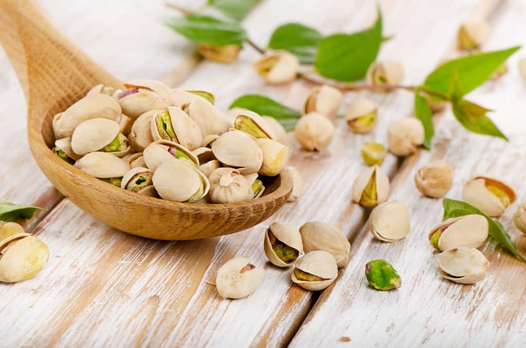 Front diagonal view of medium wooden spoon filled with pistachio nuts in the shell, with pistachios scattered with a small branch of green leaves on the white washed wooden surface.