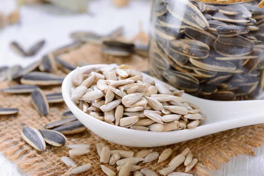 Close up diagonal view of a white ceramic spoon filled with shelled sunflower seeds next to a jar of sunflower seeds with black seed intact and both types of seeds overflowing onto a square burlap cloth on a whitewashed surface.
