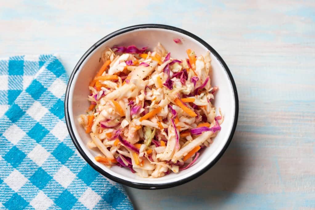 Aerial view of coleslaw with sticks of jicama in a medium white bowl with a black rim with a bright blue and white kitchen towel resting on a whitewashed light blue, rustic surface.