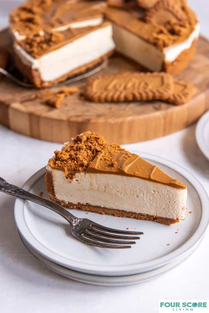 Diagonal closeup view of a slice of Biscoff cheesecake plated with a fork and a partially sliced Biscoff cheesecake placed on a round wooden cutting board in soft focus in the background. 