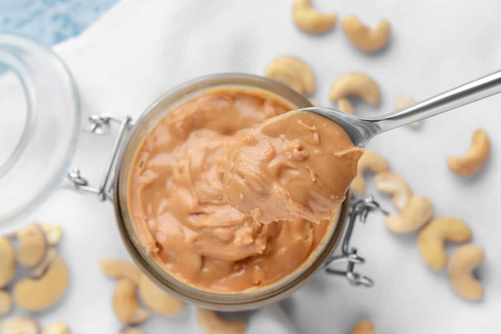 Aerial, close up view of a spoon scooping cashew butter out of a lidded jar, with cashews in soft focus on the surrounding light colored surface.