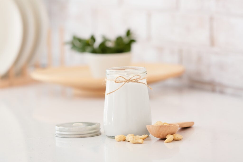Front view of a small jar of cashew milk with a natural string tied around the rim and a wooden spoon on the surface nearby overflowing with raw cashews in addition to the jar's lid on the white surface. A neutral kitchen scene is in soft focus in the background. 