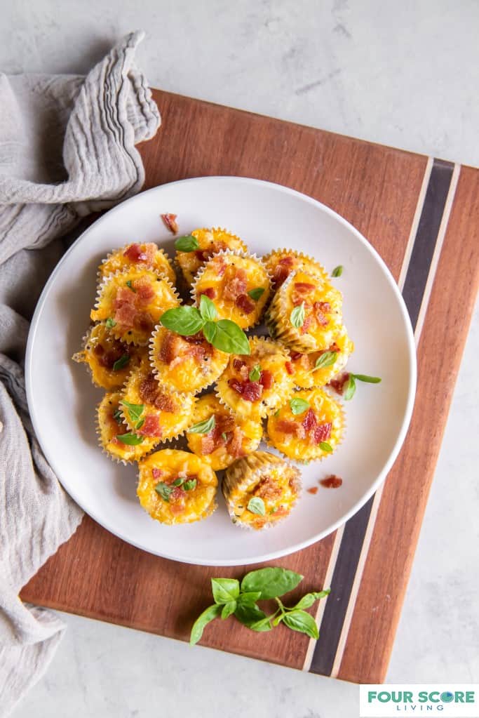 Aerial view of 12 keto egg bites piled onto a white plate with chopped bacon and fresh basil garnish, sitting on a wood cutting board with a solid light grey kitchen towel next to it, all resting on a stone surface.