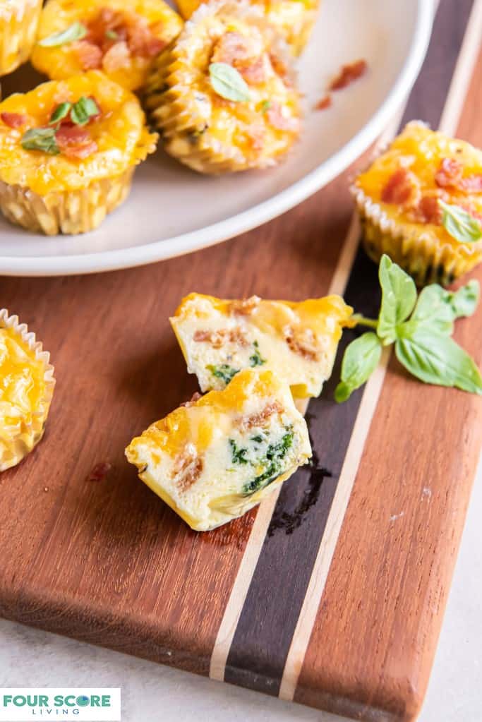 Close up Diagonal, aerial view of keto egg bites piled onto a white plate with chopped bacon and fresh basil garnish, sitting on a wood cutting board, all resting on a light colored stone surface.