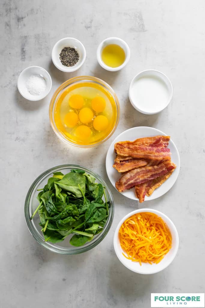 Aerial view of white and clear bowls each containing an ingredient for keto egg bites, including cooked bacon strips, shredded cheddar cheese, fresh spinach, cracked eggs, slat, pepper, oil and heavy cream all resting on a white stone surface. 