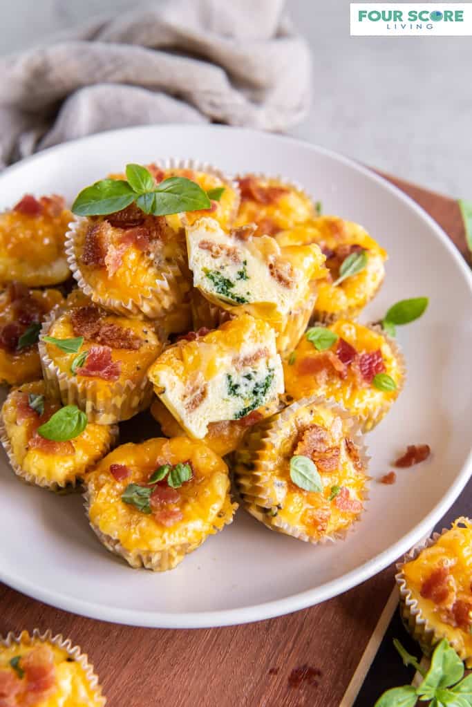 Diagonal, aerial view of egg bites piled onto a white plate with chopped bacon and fresh basil garnish, sitting on a wood cutting board with a solid light grey kitchen towel in soft focus in the background, all resting on a stone surface