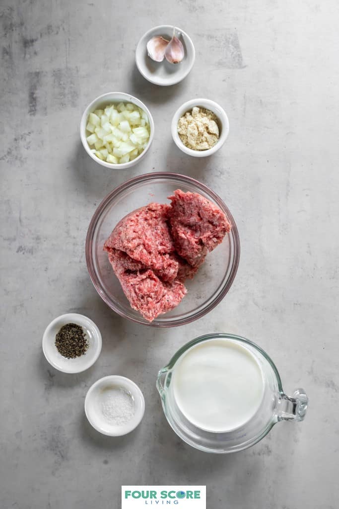 Aerial view of bowls of ingredients to make keto sausage gravy, including pork sausage without casing, salt, pepper, chopped onion, almond flour, garlic cloves, and heavy cream all resting on a white natural stone surface. 
