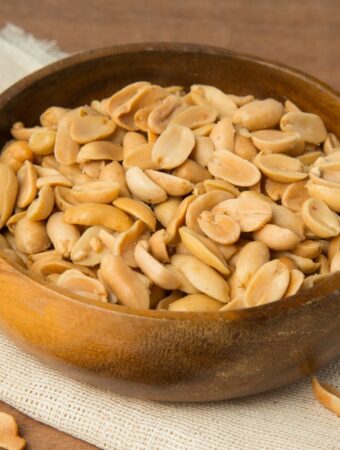 Diagonal aerial view of shelled peanuts in wooden bowl placed on la folded linen placemat on a wooden background.