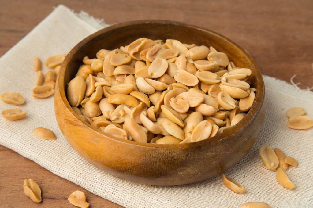 Diagonal aerial view of shelled peanuts in wooden bowl placed on la folded linen placemat on a wooden background.
