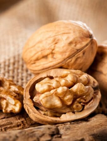 Close up frontal view of two intact walnuts in the shell and one broken open with the nut flesh exposed, one walnut half all resting on a rustic wooden surface with a burlap cloth in soft focus in the background.