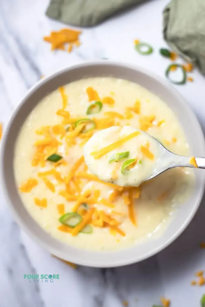 Top view photo of 4 Ingredient Potato Soup in a white bowl topped with shredded cheddar cheese and sliced green onions. There is a spoon getting a spoonful of soup.