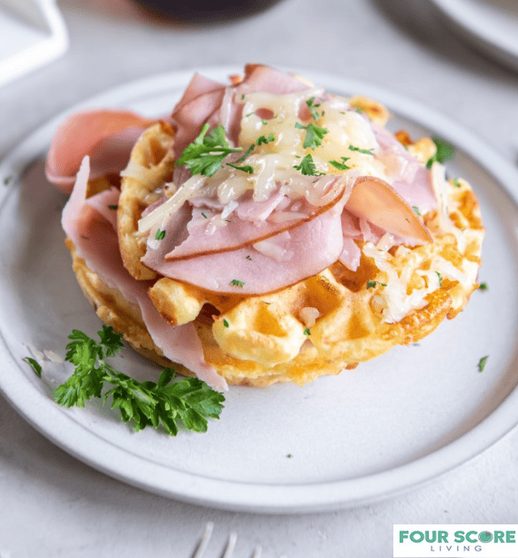 Diagonal closeup view of a keto chaffle layered with ham and melted white cheese and sprinkled with green garnish on a white plate resting on a white stone surface. 