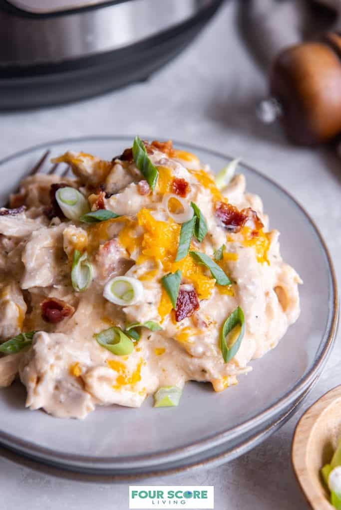 Aerial diagonal view of plated Instant Pot crack chicken dish sprinkled with chopped bacon, shredded cheddar cheese and green scallions with an Instant Pot and a wooden pepper grinder in partial view and soft focus in the background.