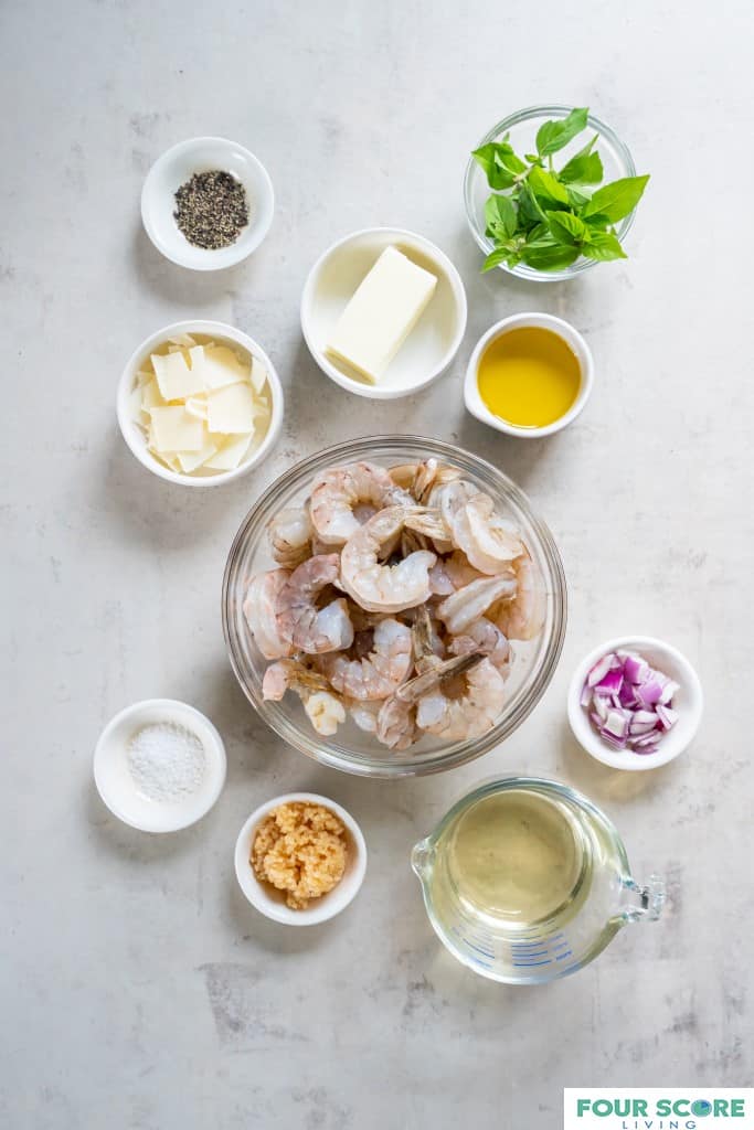 Aerial view of small and medium bowls holding the ingredients to make keto shrimp scampi, including avocado oil, a half stick of butter, slices of fresh Parmesan cheese, raw shrimp with tails on, chopped red onion, minced garlic, salt and white wine. 