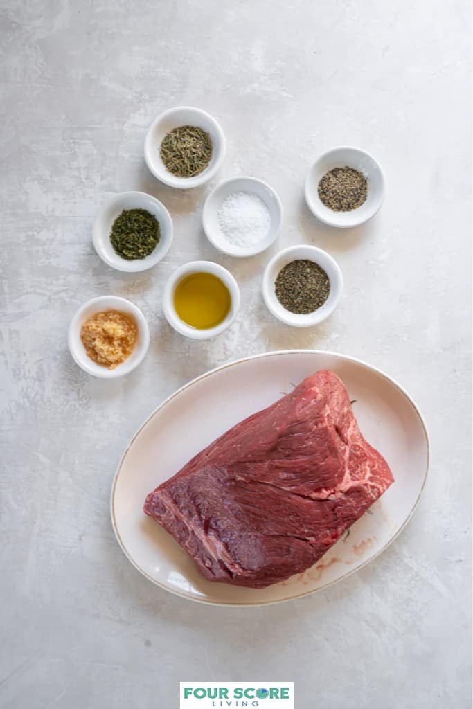 Aerial view of small white dishes and an oval white platter with the ingredients to make an herb crusted bottom round roast, including, a bottom round roast, olive oil, garlic, herbs, salt and pepper.