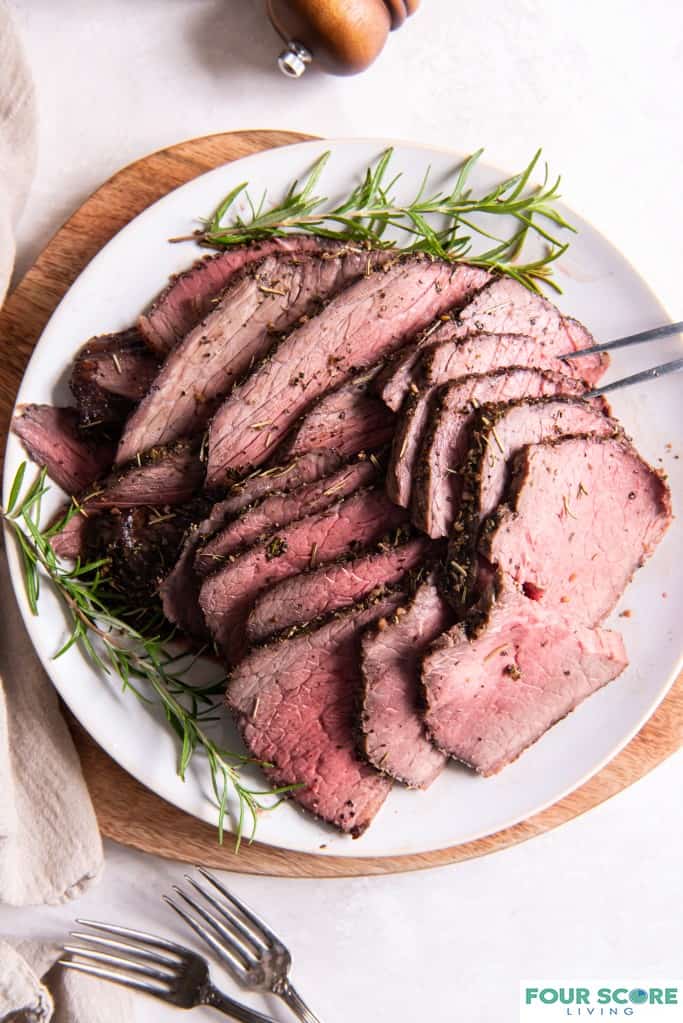 Aerial view of cooked and sliced herb crusted beef bottom round roast arranged on a white plate with sprigs of fresh rosemary layered on a round wooden cutting board  with forks, a natural colored kitchen towel and a wooden pepper mill in partial view, all resting on a white stone surface. 