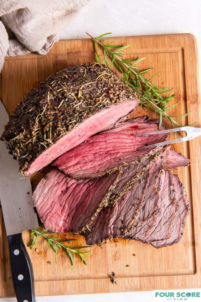 Aerial view of herb crusted bottom round roast partially sliced on a wood cutting board with a spring of fresh rosemary and a two pronged serving tool and a sharp knife and a natural cloth kitchen towel all resting on a white stone surface.
