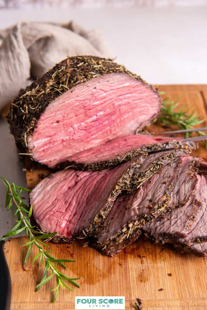 Diagonal view of a cooked and partially sliced bottom round roast on a wooden cutting board with sprigs of fresh rosemary. 