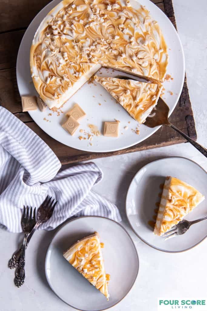 Aerial view of a caramilk cheesecake on a white platter, resting on top of a dark brown wooden cutting board, with 2 slices served onto white plates and a pinstriped kitchen towel and forks, all resting on a white stone surface.