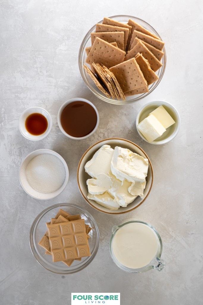 Aerial view of bowls and dishes filled with the ingredients to make caramilk cheesecake, including, cream cheese, heavy cream, unsalted butter, graham crackers, caramilk chocolate bars, granulated sugar, caramel sauce, vanilla extract and cream cheese.