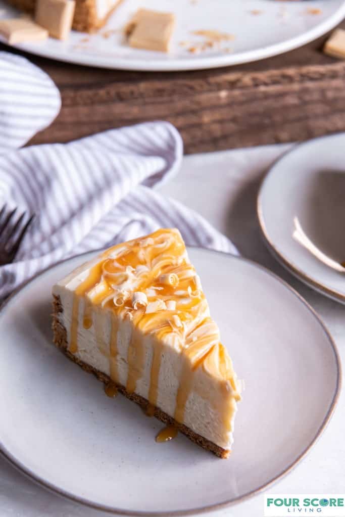 Diagonal up close view of a single slice of caramilk cheesecake on a white plate, garnish with melted caramel and chocolate shavings with a a pinstripe kitchen towel, fork and a partial view of a plate holding a cut caramilk cheesecake in soft focus in the background. 