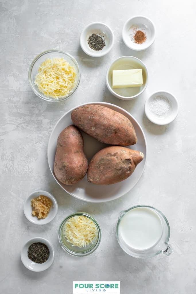Aerial view of white bowls and clear glass bowls filled with the ingredients to make scalloped sweet potatoes, including 3 raw sweet potatoes, cheese, salt, pepper, butter, minced garlic, dried thyme, nutmeg, heavy cream and gruyere cheese.