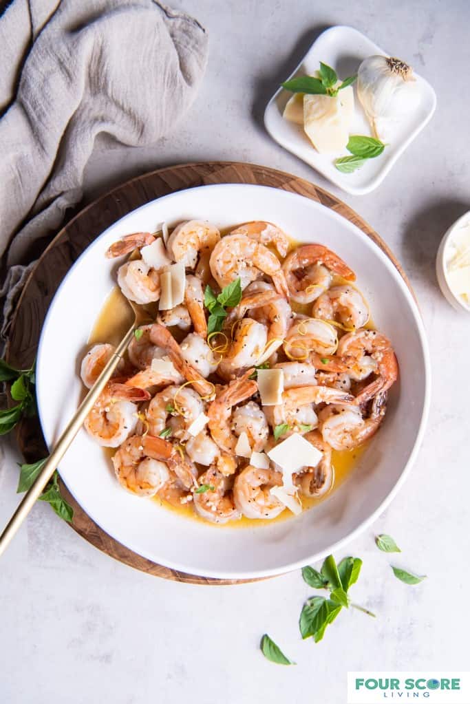 Aerial view of a white white bowl filled with keto shrimp scampi in a white wine garlic butter sauce garnished with fresh basil leaves and sliced pieces of fresh Parmesan cheese placed on a wooden cutting board with a small shite dish holding a fresh garlic bulb and small blocks of Parmesan cheese with a light grey natural fiber dish towel all resting on a white stone surface.