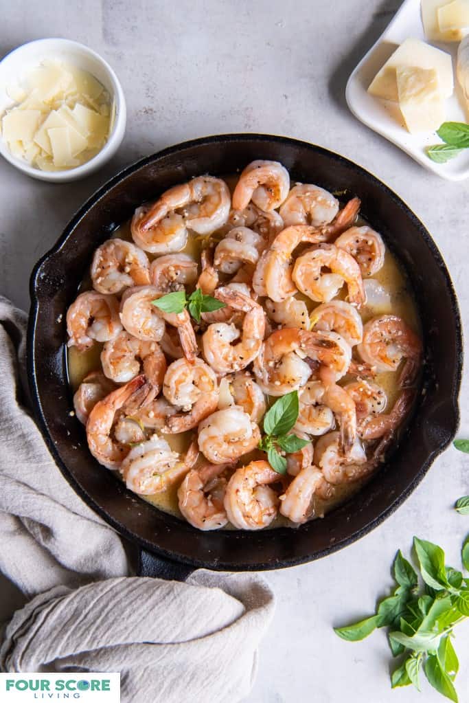 Aerial view of a cast iron skillet filled with prepared keto shrimp scampi in a white wine garlic butter sauce garnished with fresh basil leaves with a small shite dish holding a fresh garlic bulb and small blocks of Parmesan cheese and a small bowl of sliced fresh Parmesan cheese with a light grey natural fiber dish towel all resting on a white stone surface.