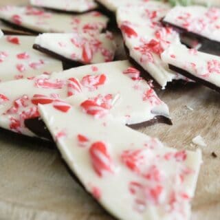 Diagonal view of peppermint bark, made with a layer of semisweet chocolate, white chocolate and crushed candy cane on a round light colored wooden cutting board with a sprig of fresh rosemary in soft focus in the background, all resting on a white stone surface.