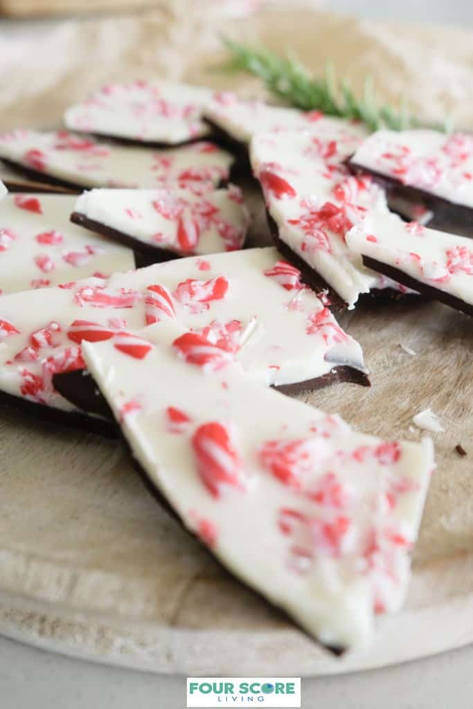 Diagonal view of peppermint bark, made with a layer of semisweet chocolate, white chocolate and crushed candy cane on a round light colored wooden cutting board with a sprig of fresh rosemary in soft focus in the background, all resting on a white stone surface.