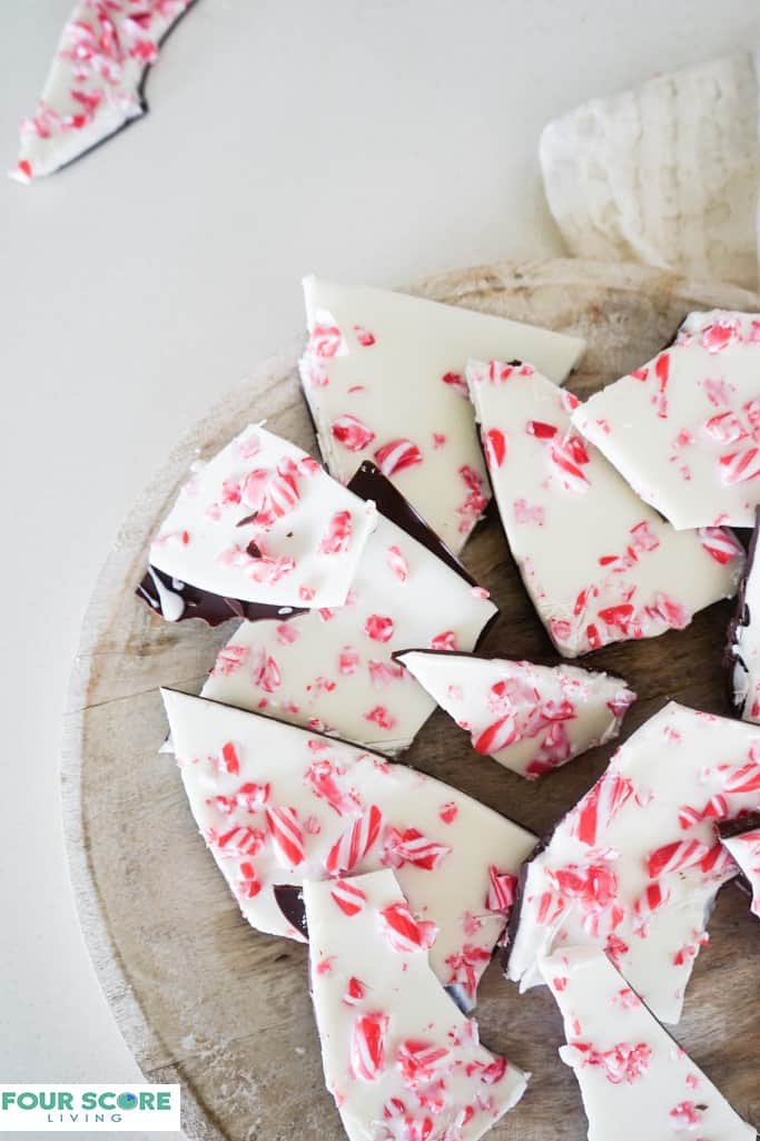 Close up aerial view of peppermint bark, made with a layer of semisweet chocolate, white chocolate and crushed candy cane on a round light colored wooden cutting board all resting on a white stone surface.