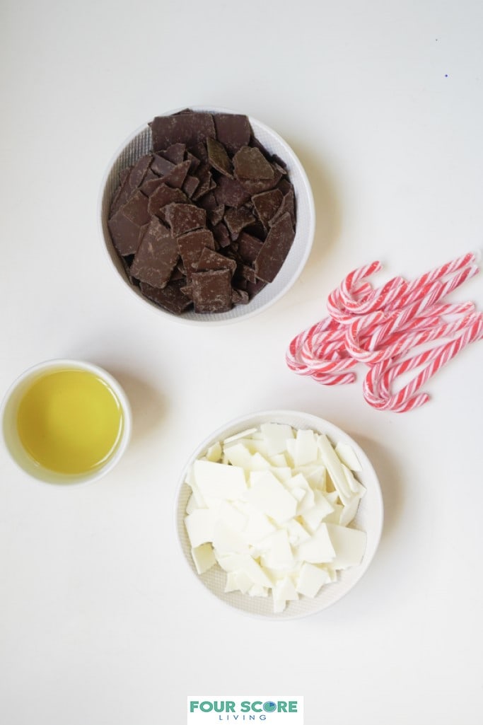 Aerial view of small white bowls holding the ingredients to make peppermint bark, including full size candy canes, pieces of white chocolate, pieces of semisweet chocolate and oil.