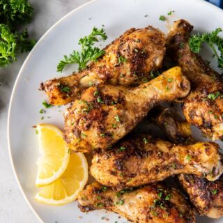 Baked chicken legs plated on a white plate with lemons slices and chopped fresh herb garnish all resting on a white stone surface.