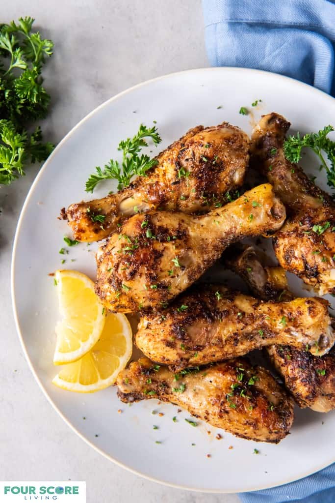 Aerial view of prepared baked chicken legs plated on a white plate with lemons slices and chopped fresh herb garnish all resting on a white stone surface. 