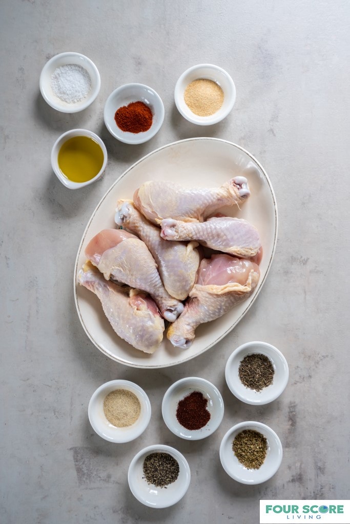 Aerial view of ingredients to make baked chicken legs, including 6 uncooked chicken legs on an oval white platter and small white dishes of herbs and spices.