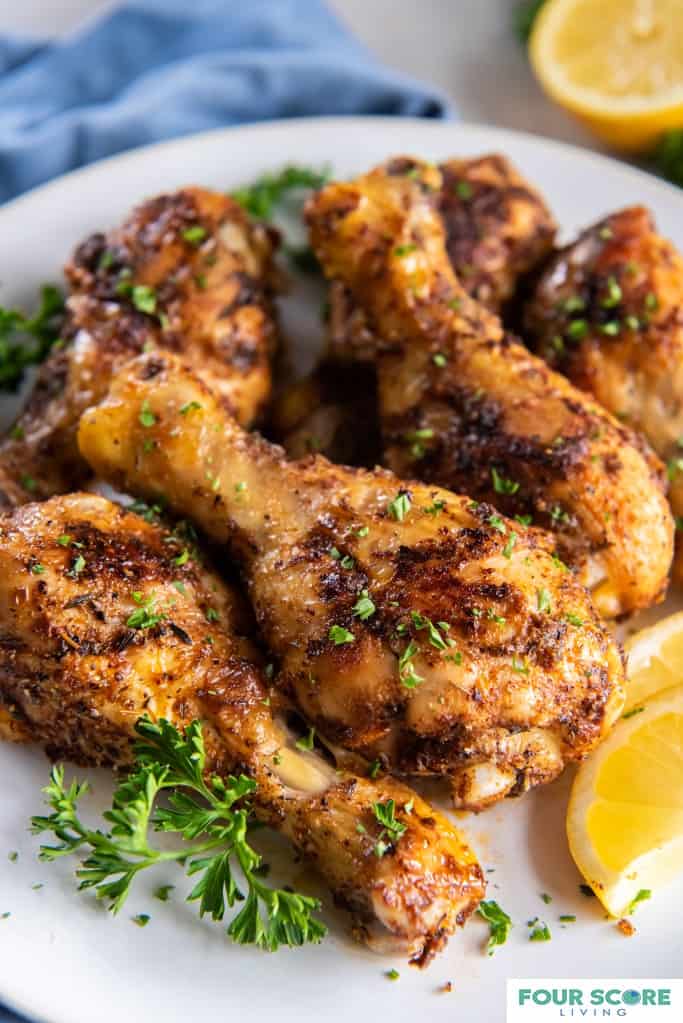 Close up diagonal view of seasoned and cooked baked chicken legs, plated on a white plate with lemons slices and chopped fresh herb garnish. 